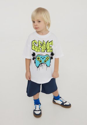 T-shirt bianca con una grafica audace "GAME" in giallo neon e un design di un controller di gioco blu, abbinata a pantaloni corti blu scuro e sneakers multicolori.
