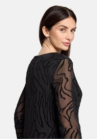 Czarny top z teksturowanym wzorem z uniesionymi, falistymi detalami. Rękawy są przezroczyste, ukazując misterny wzór na solidnej podstawie.