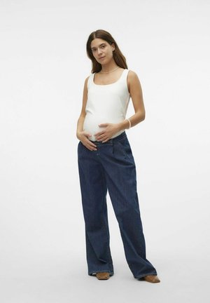 White tank top with a square neckline, paired with loose-fitting dark blue denim pants. Model has long brown hair and is standing with hands on belly.