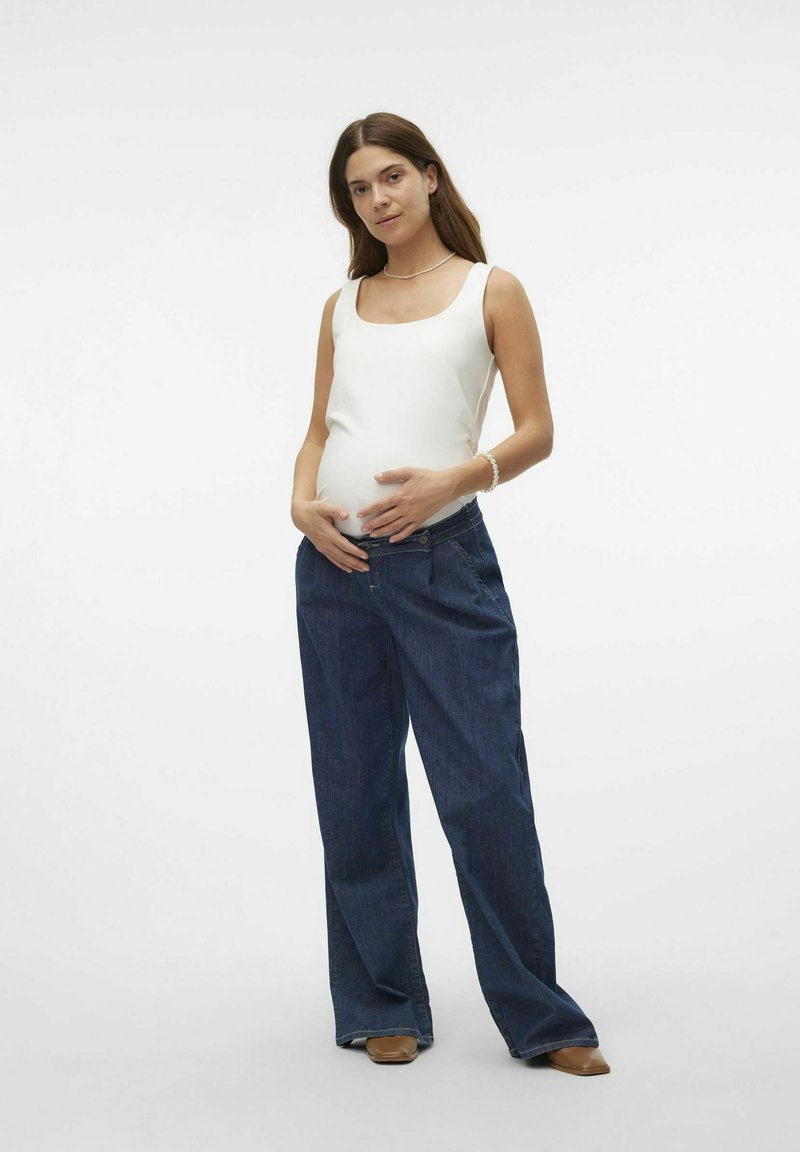 Un débardeur blanc à décolleté carré, associé à un pantalon en denim bleu foncé ample. Le mannequin a de longs cheveux bruns et se tient les mains sur le ventre.