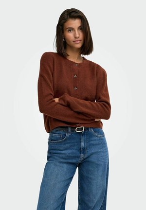 Femme aux cheveux raides et foncés portant un pull marron boutonné et un jean bleu avec une ceinture noire, debout les bras croisés.