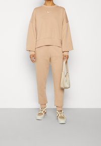 Beige mikina Nike s kleslými rameny, spárovaná s odpovídajícími zúženými tepláky. Doplňky zahrnují světle barevnou tašku a tenisky.