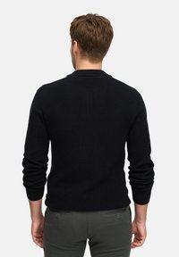 Schwarzer Strickpullover mit langen Ärmeln und gerippter Textur. Der Rücken hat einen Rundhalsausschnitt und enganliegende Bündchen. Getragen mit dunkelgrauen Hosen.