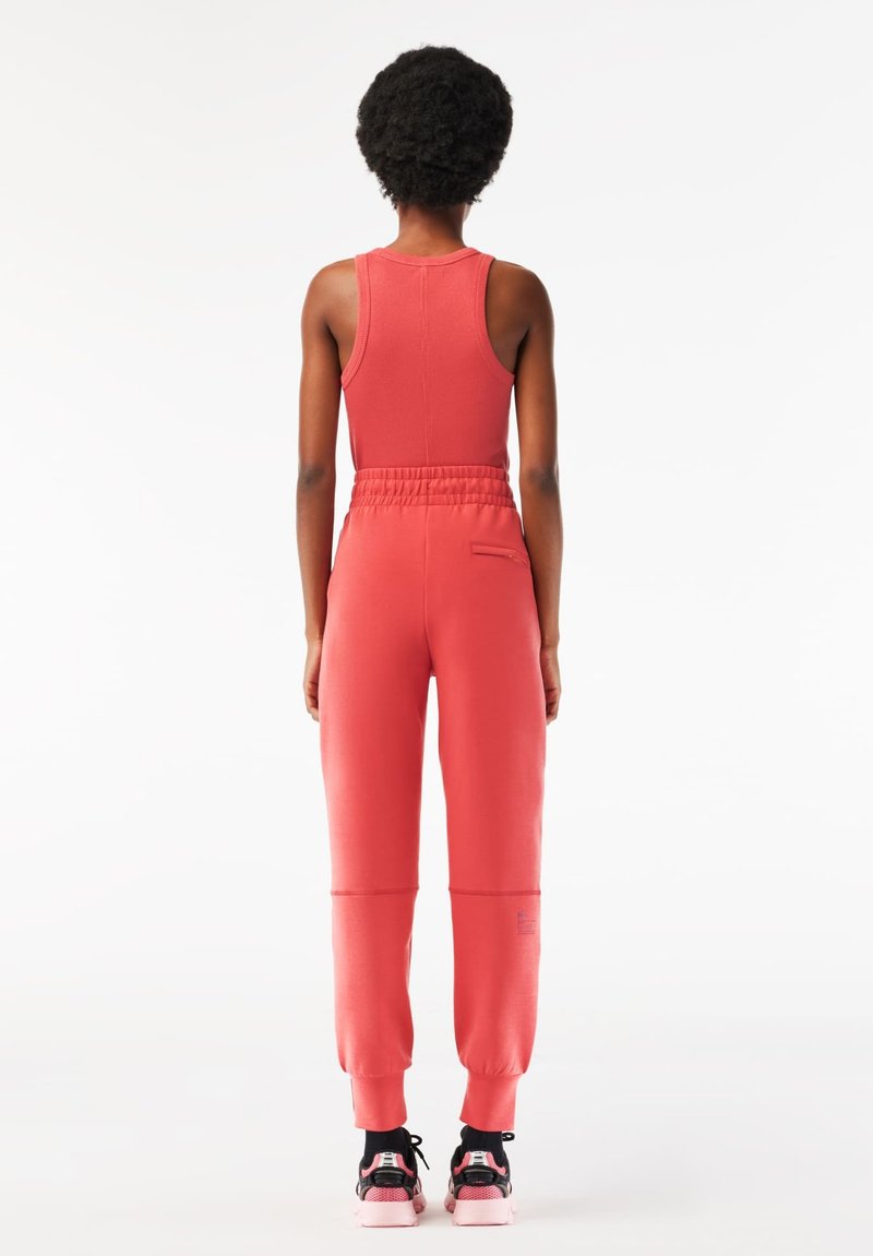 Korallenfarbenes Sportoutfit mit einem ärmellosen Top und schmal zulaufenden Jogginghosen mit elastischem Bund. Strukturiertes Material und kontrastierende Schuhe.