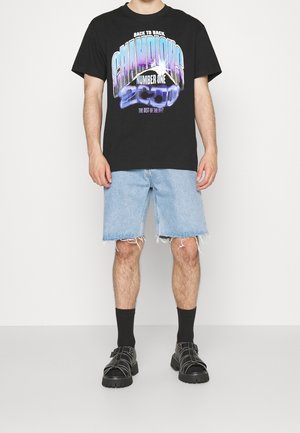 Zwart grafisch T-shirt met "CHAMPIONS 2000" opdruk, gecombineerd met losse lichtblauwe denimshorts met rafelige zoom en zwarte sandalen.