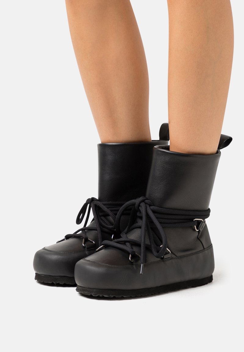 Jambes chaussées de bottines en cuir noir à semelles épaisses et lacets noirs enroulés autour des chevilles, présentées sur un fond blanc.