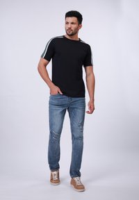 Camiseta de algodón negra con acentos en verde y blanco a rayas en los hombros, combinada con jeans claros desgastados y zapatillas color tostado.