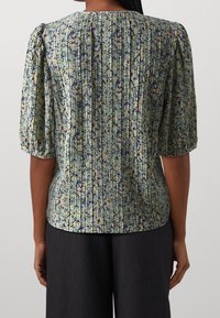 Blusa texturizada en verde con patrones florales en amarillo y púrpura. Cuenta con mangas cortas abullonadas y una cintura ajustada con diseño plisado.