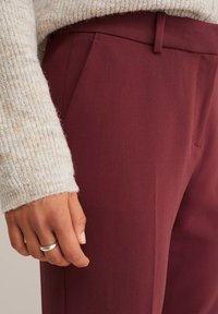 Burgundybyxor med en slät yta och en skräddarsydd passform, som har sidofickor och en matchande midjeresår, kompletterade med en ljus tröja.