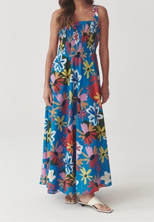 Vrouw in een blauwe mouwloze maxi-jurk met grote meerkleurige bloemenprints en witte open hiel sandalen.