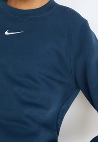 Námořnická modrá mikina vyrobená z měkké látky, s kulatým výstřihem a bílým logem Nike na hrudi. Hladký povrch s detaily švů.