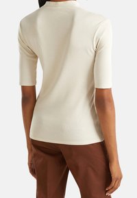 Beige kortärmad topp med hög hals, tillverkad av mjukt tyg. Har en figurnära silhuett och subtil sömdetaljering vid fållen.