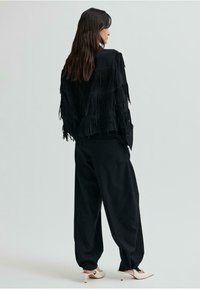 Čierna nubuková bunda s strapcami, kombinovaná s voľnými čiernymi nohavicami a bielymi topánkami na podpätku so šnúrkami okolo členkov, pohľad zozadu.