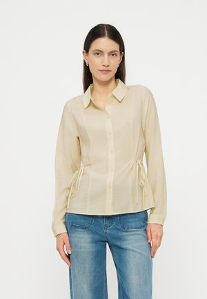 Frau mit langen dunklen Haaren, die ein beiges Hemd mit Knöpfen und seitlichen Bändern sowie blaue Jeans trägt, steht vor einem schlichten hellen Hintergrund.