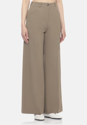 Donna con pantaloni beige a vita alta e gamba larga e scarpe bianche, in piedi con le mani rilassate lungo i fianchi davanti a uno sfondo semplice.