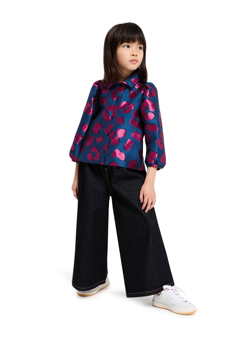Blouse en satin floral bleu avec des motifs abstraits roses, devant à bouton et manches bouffantes, associée à un pantalon en denim large et foncé et des baskets blanches.