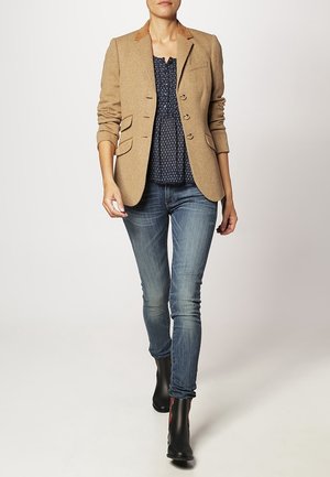 Femme marchant, portant un blazer beige aux manches retroussées, un chemisier sombre à motifs, un jean bleu et des bottines noires, sur un fond uni.
