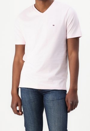 Person trägt ein hellrosa V-Ausschnitt-T-Shirt mit kurzen Ärmeln und kleinem Logo auf der Brust sowie blaue Jeans aus Denim vor weißem Hintergrund.