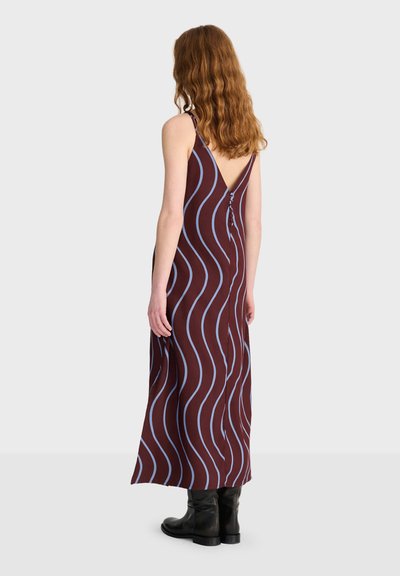 Femme aux cheveux ondulés portant une robe longue marron sans manches avec des rayures ondulées bleues, des boutons dans le dos et des bottines noires, debout de dos.