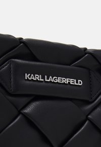 Svart läderhandväska med en texturerad, quiltad design och en metallplatta med logotypen "Karl Lagerfeld." Har släta och upphöjda detaljer.