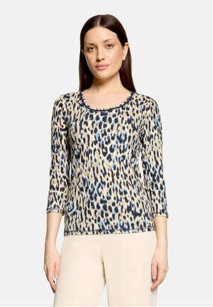 Vrouw met steil donker haar draagt een beige, zwarte en blauwe luipaardprint lange mouwen top en lichtbeige broek tegen een neutrale achtergrond.
