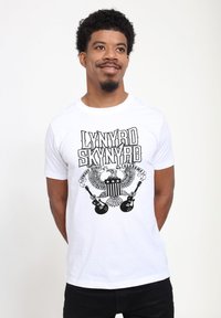 Bílý bavlněný tričko s černou grafikou Lynyrd Skynyrd, včetně orla, hvězd a kytar, s krátkými rukávy a kulatým výstřihem.