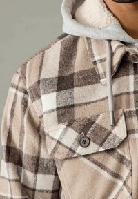 Beżowa i brązowa kraciasta kurtka z miękką, poliestrową podszewką i szarym kapturem. Posiada dwie kieszenie na piersi i brązowe guziki. Miękka tekstura i ciepły design.