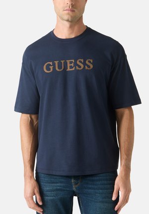 Mężczyzna w granatowym T-shircie z krótkim rękawem z dużym brązowym logo "GUESS" na klatce piersiowej i niebieskich dżinsach, stojący na jednolitym tle.