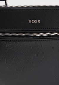 Μαύρη δερμάτινη τσάντα χειρός με υφή, που διαθέτει ασημένιο φερμουάρ και το ανάγλυφο λογότυπο "BOSS" στην πρόσοψη.
