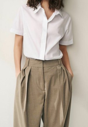 Personne portant une chemise blanche à manches courtes boutonnée, rentrée dans un pantalon plissé taille haute beige à fines rayures, les mains dans les poches.