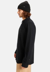 Zwarte, knoopsluiting shirt gemaakt van zachte stof, met een zak aan de linkerkant, lange mouwen en knoopmanchetten. Gecombineerd met een zwarte gebreide beanie.