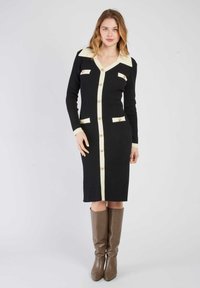 Czarna dzianinowa sukienka z kremowymi akcentami, dekoltem w serek, długimi rękawami, z zapięciem na guziki i przednimi kieszeniami. Nosi się ją z brązowymi, wysokimi na kolana botkami.