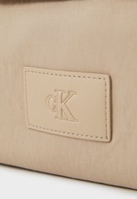 Borsa in tessuto beige con una superficie liscia, dotata di un'etichetta in pelle rettangolare impressa con "cK" e bordi cuciti.