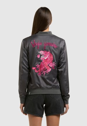 Donna che indossa una giacca bomber in raso nero con una tigre ricamata in rosa brillante e la scritta "khujo genuine" sul retro, abbinata a pantaloncini di denim neri.