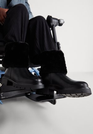 Czarne skórzane botki z grubą, teksturowaną podeszwą i puszystymi czarnymi futrzanymi mankietami. Są umieszczone na podnóżku wózka inwalidzkiego.