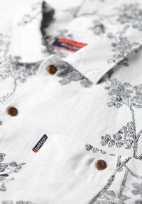 Camisa de manga corta blanca con un patrón floral negro. Botones marrones, bolsillo en el pecho y etiqueta con el logo "Superdry" visible. Material de algodón.