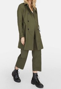 Olijfgroene trenchcoat met een dubbelgeknoopte ontwerp, zwarte knoppen, gecombineerd met bijpassende cropped broek en zwarte veterschoenen.