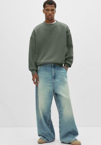 Zöld, oversize pulóver hosszú ujjakkal, amelyet széles lábú, fakó kék farmernadrággal viselnek. A modell egy sima fehér háttér előtt áll.