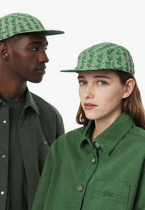 Groen gepatternde pet met een platte rand, voorzien van een geometrisch ontwerp. De modellen dragen bijpassende groene outfits, waardoor de details van de pet benadrukt worden.