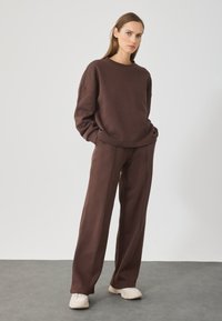 Brązowy bawełniany sweatshirt i dopasowane szerokie spodnie. Sweatshirt ma okrągły dekolt i opadające ramiona; spodnie mają boczne szwy i kieszenie.