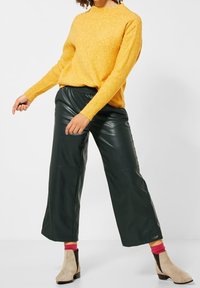 Gul ribbad tröja parat med mörkgröna byxor i konstläder med vida ben. Modellen bär beige ankelstövlar med svarta detaljer och rosa strumpor.