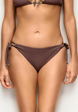 Kobieta nosząca brązowe teksturowane dół bikini z wiązaniami po bokach, ukazująca środkową część ciała i górne partie ud na jednolitym tle.