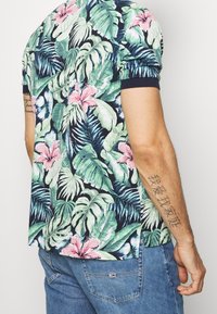 Koszulka polo w kwiaty z zielonymi liśćmi i różowymi kwiatami, krótkimi rękawami, granatowym kołnierzem i rozcięciami po bokach. Noszona z niebieskimi jeansami.