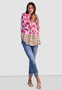 Langärmlige Bluse in pink-beigen Mustern, mit V-Ausschnitt und lockerer Passform, kombiniert mit hellblauen Jeans mit umgeschlagenen Säumen und Sandalen.