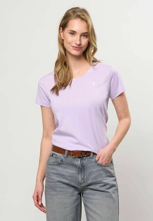Ung kvinde med blondt hår iført en lyse lilla t-shirt, stukket ned i højtaljede grå jeans med et brunt bælte, hånd i lommen.