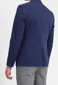 Man som bär en figursydd marinblå kavaj med knäppta manschetter och grå strukturerade byxor, sedd bakifrån mot en vit bakgrund.