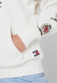 Sweatshirt bianco con crest ricamato sulla manica, polsini a coste e un patch con il logo Tommy Jeans sul fianco. Materiale in cotone morbido.