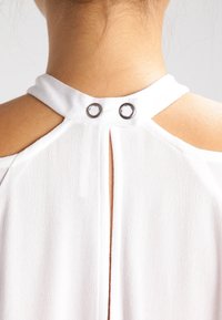 Vit ärmlös topp med design av nyckelhål baktill, prydd med metalliska detaljer och en texturerad tyg. Detaljer vid halsringningen och axelsnitt.