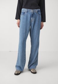 Světle modré džíny s volným střihem, vybavené knoflíkovým zapínáním, pěti kapsami a hladkým povrchem. Nosené s vzorovanými botami.