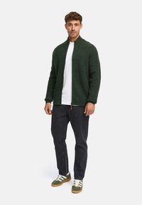 Grüne Strickjacke mit Reißverschluss und hohem Kragen, getragen über einem weißen Hemd, kombiniert mit dunklen Jeans und grünen Sneakers mit weißen Streifen.